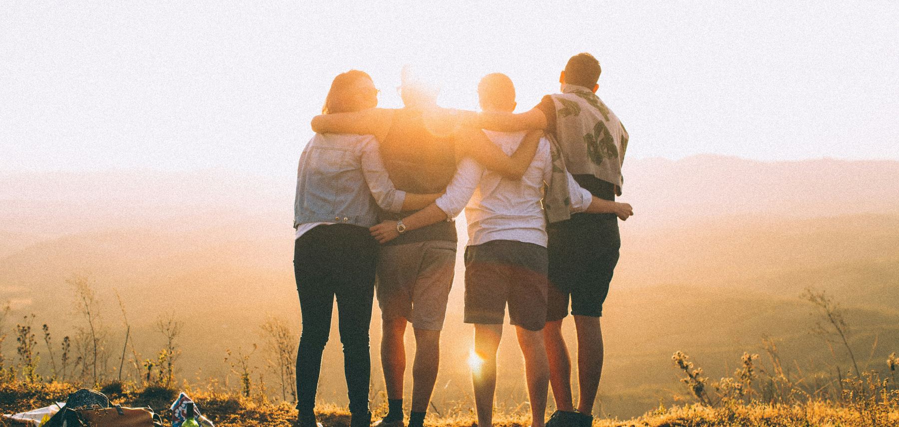 Four friends with their arms around each other, watching the sunset over hills