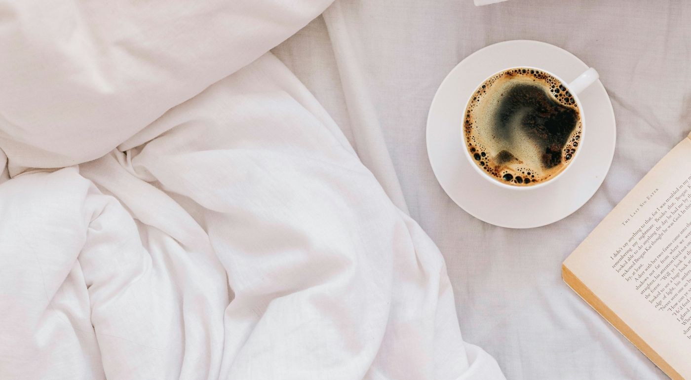 A cup of black coffee and an open book on a white duvet