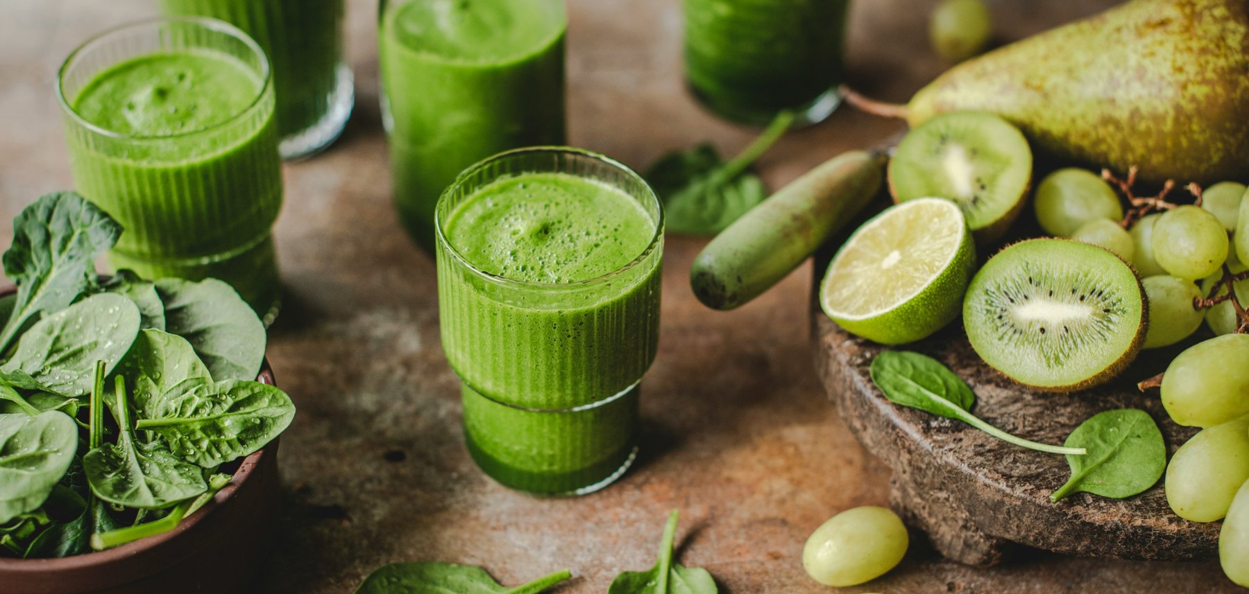 Glasses of green smoothie beside spinach, kiwi, lime, pears and green grapes on a rustic table