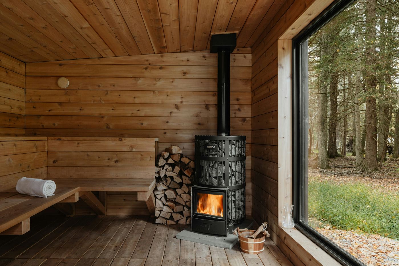A wood-paneled sauna with a wood stove and a large window onto a forest